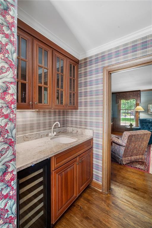 bar with dark wood-type flooring, sink, crown molding, light stone counters, and beverage cooler