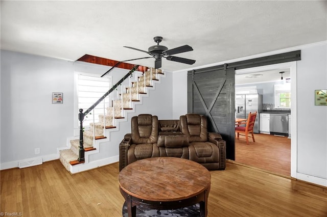 living area with a barn door, stairway, light wood-style flooring, ceiling fan, and ornamental molding