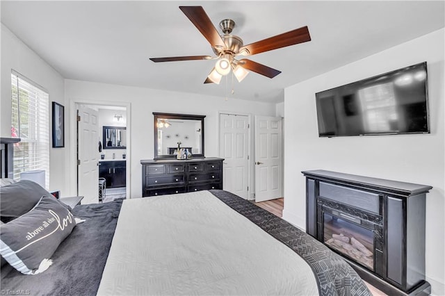 bedroom featuring a glass covered fireplace, a ceiling fan, ensuite bath, and wood finished floors