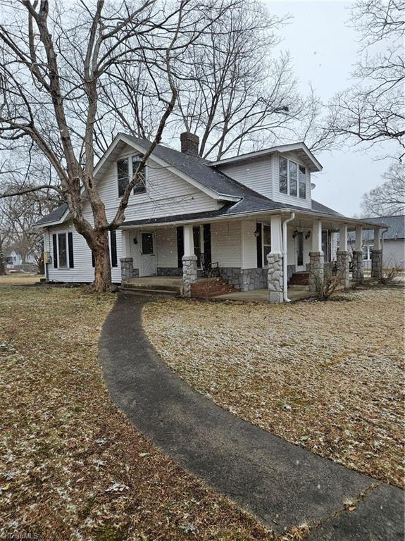 view of front facade featuring covered porch, stone siding, a chimney, and roof with shingles