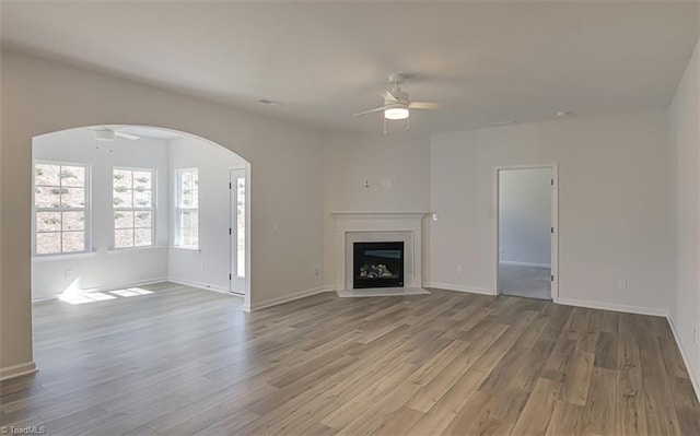 unfurnished living room with a fireplace with flush hearth, arched walkways, ceiling fan, and wood finished floors