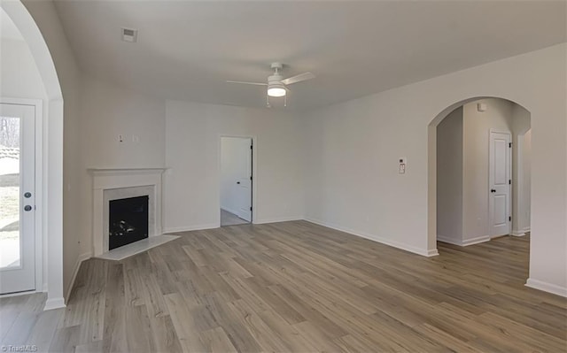 unfurnished living room with arched walkways, a fireplace, light wood finished floors, visible vents, and a ceiling fan