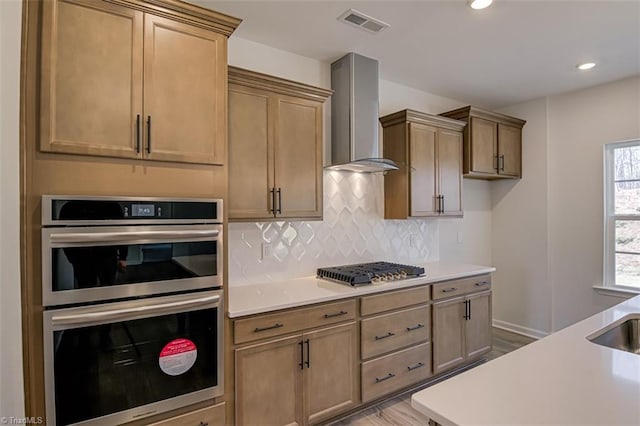 kitchen with stainless steel appliances, wall chimney exhaust hood, backsplash, and light countertops