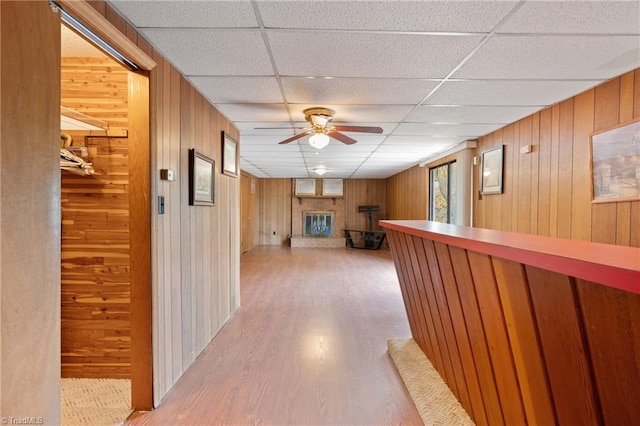 hall with light wood-type flooring, a drop ceiling, and wooden walls