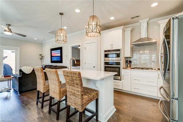 kitchen with wall chimney exhaust hood, white cabinetry, decorative light fixtures, stainless steel appliances, and a kitchen island with sink