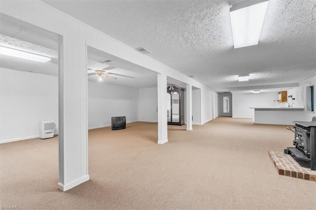 basement featuring ceiling fan, light colored carpet, a wood stove, and a textured ceiling