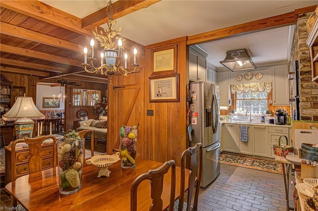 dining space featuring sink, beamed ceiling, ceiling fan with notable chandelier, and wooden walls