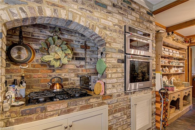 kitchen with white cabinetry, stainless steel double oven, brick wall, gas stovetop, and beamed ceiling