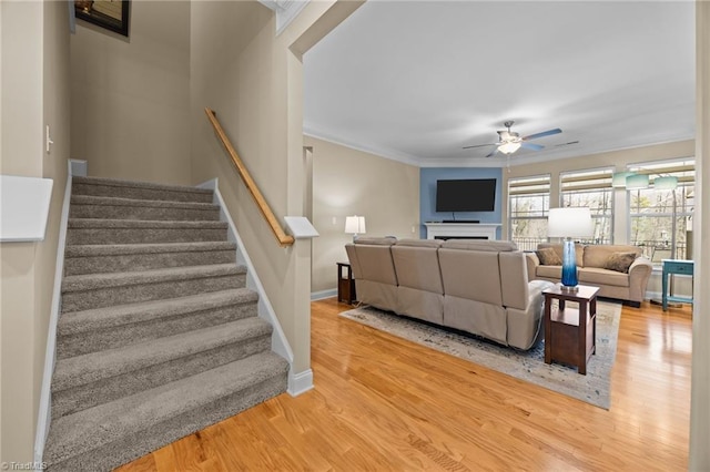 living room featuring a ceiling fan, crown molding, light wood finished floors, baseboards, and stairs