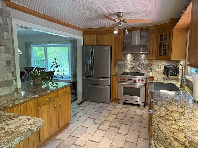 kitchen featuring appliances with stainless steel finishes, wall chimney exhaust hood, brown cabinets, backsplash, and a ceiling fan