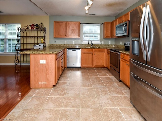 kitchen with light stone counters, stainless steel appliances, a sink, a peninsula, and brown cabinetry