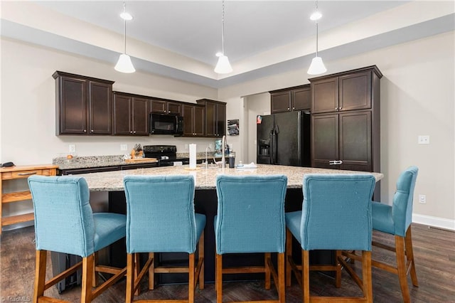 kitchen featuring dark brown cabinetry, a kitchen island with sink, dark wood finished floors, light stone counters, and black appliances