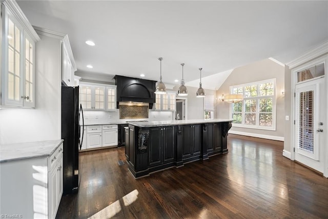 kitchen with black refrigerator, hanging light fixtures, custom range hood, white cabinets, and an island with sink