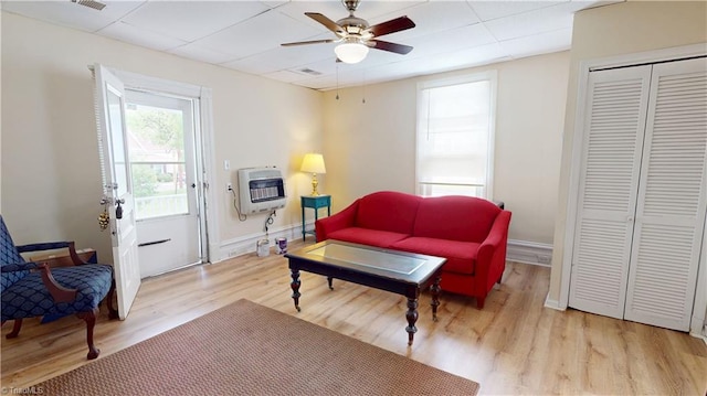 living area featuring a ceiling fan, heating unit, light wood-style flooring, a drop ceiling, and baseboards
