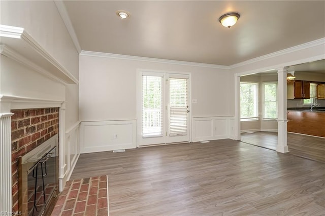 unfurnished living room featuring crown molding, a brick fireplace, wainscoting, decorative columns, and wood finished floors