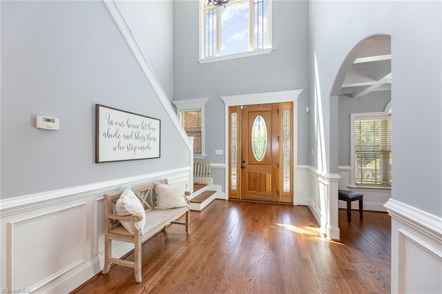 foyer with arched walkways, a towering ceiling, wood finished floors, and a wainscoted wall