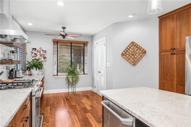 kitchen with wood finished floors, appliances with stainless steel finishes, brown cabinetry, wall chimney range hood, and baseboards