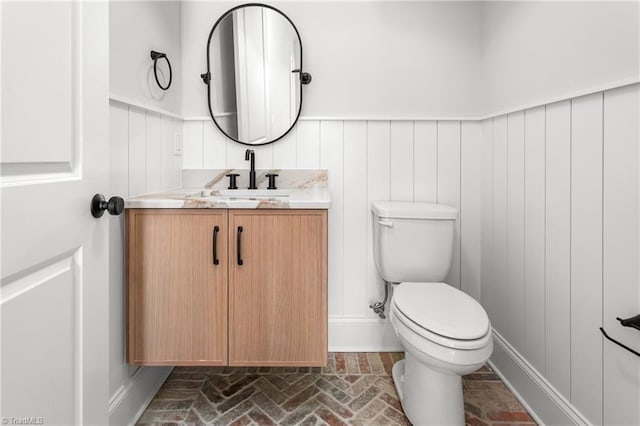 bathroom featuring wainscoting, toilet, vanity, and brick floor