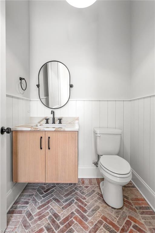 bathroom with a wainscoted wall, toilet, vanity, and brick floor