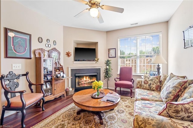 living area featuring a warm lit fireplace, ceiling fan, visible vents, and wood finished floors