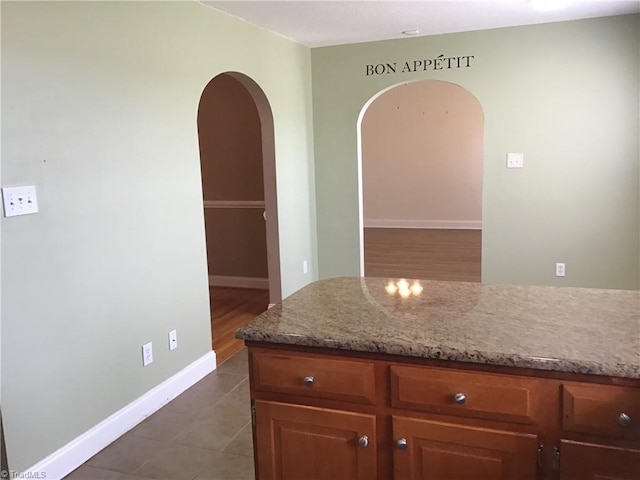 kitchen with arched walkways, brown cabinets, light stone countertops, dark tile patterned flooring, and baseboards