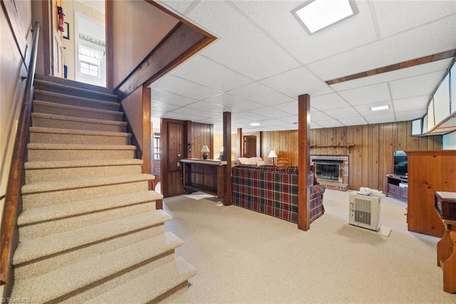 basement featuring a drop ceiling, wood walls, carpet flooring, a brick fireplace, and stairway