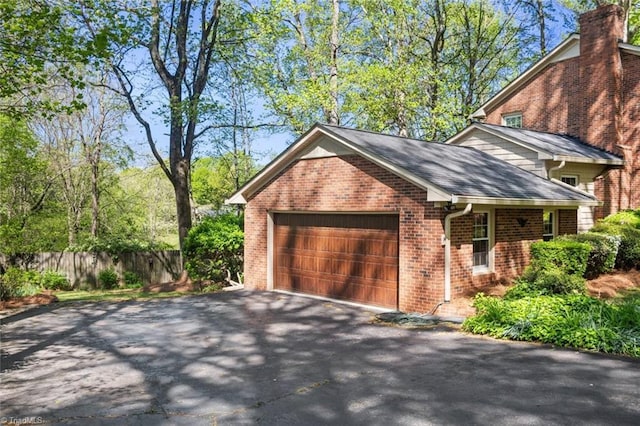 exterior space featuring aphalt driveway, a chimney, an attached garage, fence, and brick siding