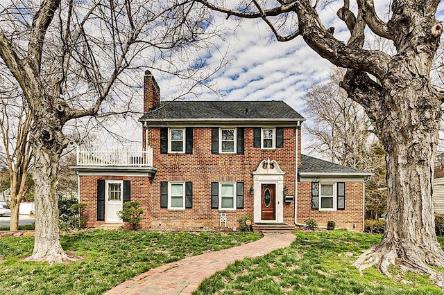 colonial home with brick siding, a chimney, a front yard, and a balcony