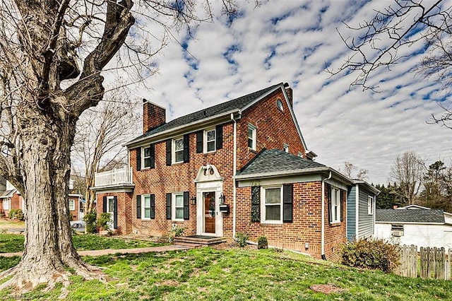 colonial inspired home featuring a balcony, brick siding, fence, a chimney, and a front yard