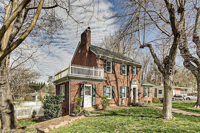 view of front facade featuring brick siding, a chimney, a front yard, fence, and a balcony