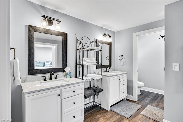 ensuite bathroom with baseboards, toilet, two vanities, wood finished floors, and a sink