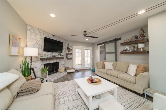 living room featuring a ceiling fan, wood finished floors, a stone fireplace, a barn door, and recessed lighting