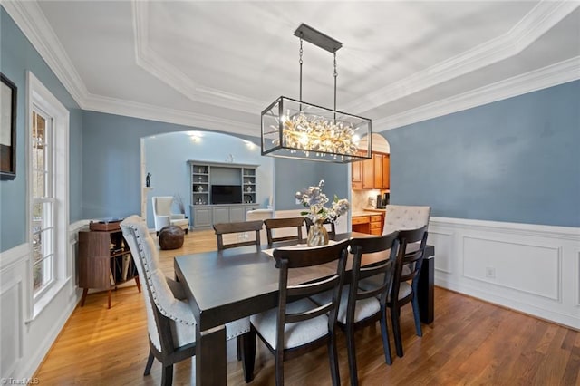 dining room with arched walkways, a raised ceiling, wainscoting, and light wood finished floors