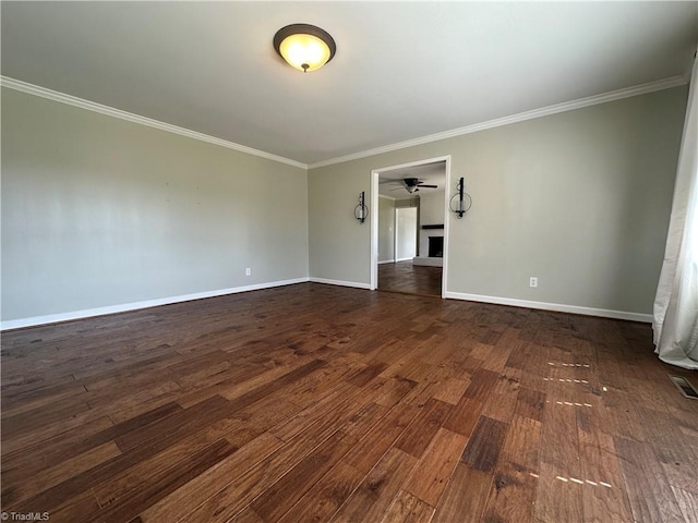 spare room featuring a fireplace, a ceiling fan, baseboards, dark wood-style floors, and crown molding