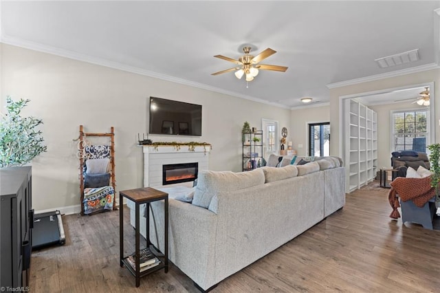 living area with visible vents, a ceiling fan, a glass covered fireplace, ornamental molding, and wood finished floors