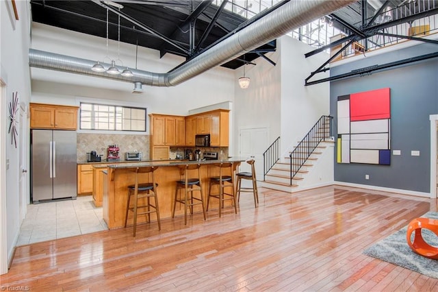 kitchen with light hardwood / wood-style floors, tasteful backsplash, stainless steel refrigerator, a kitchen breakfast bar, and a towering ceiling