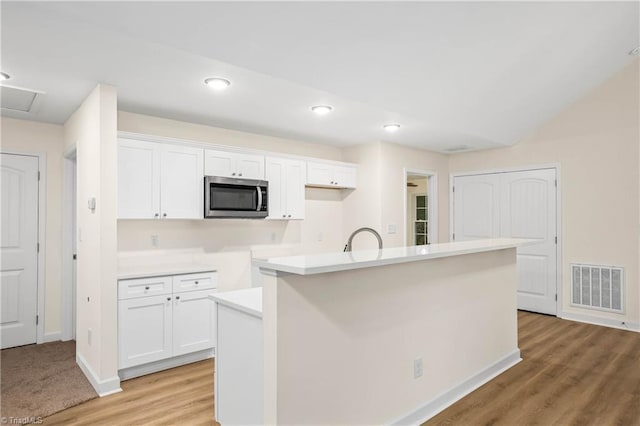 kitchen featuring visible vents, an island with sink, white cabinets, stainless steel microwave, and light wood-type flooring