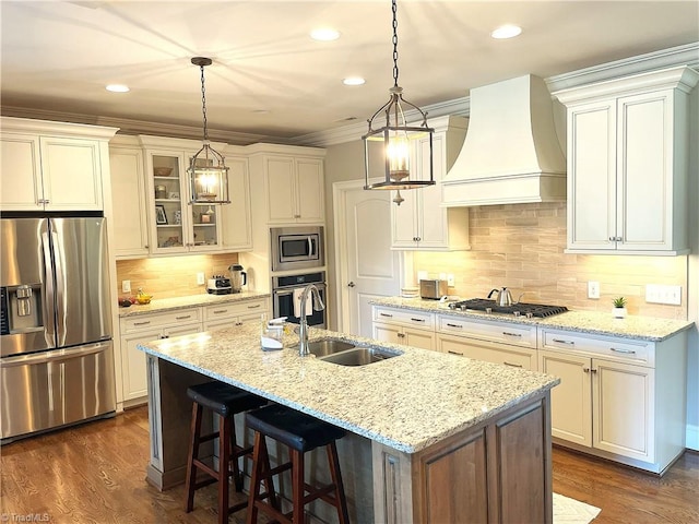 kitchen with a center island with sink, a sink, stainless steel appliances, glass insert cabinets, and custom exhaust hood