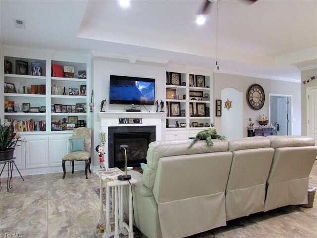 living room featuring ornamental molding, a fireplace, a tray ceiling, built in features, and ceiling fan