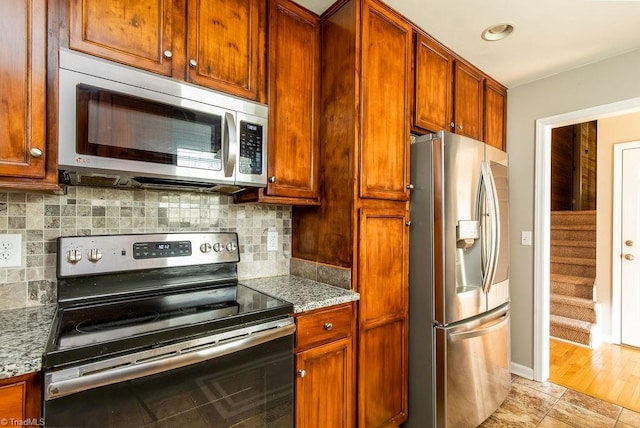 kitchen featuring light stone counters, stainless steel appliances, decorative backsplash, brown cabinetry, and baseboards