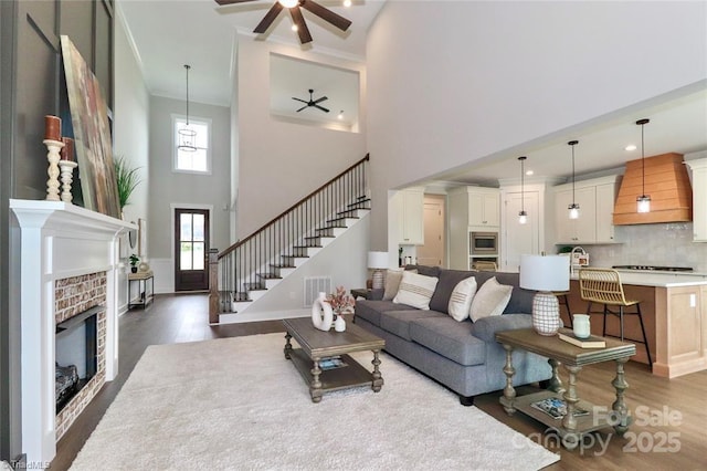 living area with stairway, a ceiling fan, a brick fireplace, dark wood finished floors, and visible vents