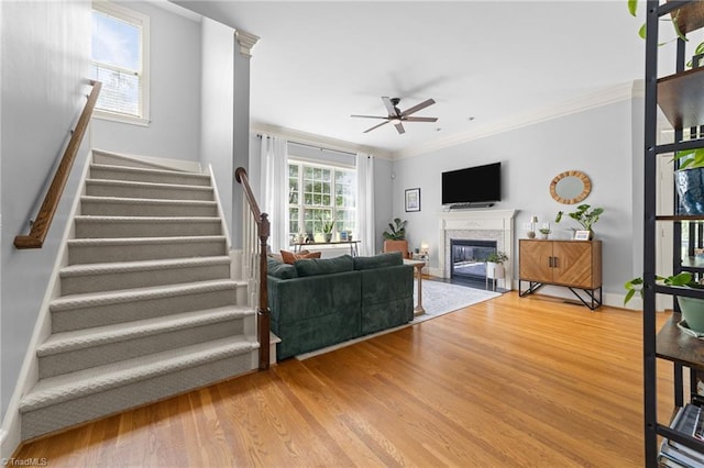 living room with ornamental molding, stairs, a fireplace with flush hearth, wood finished floors, and a ceiling fan