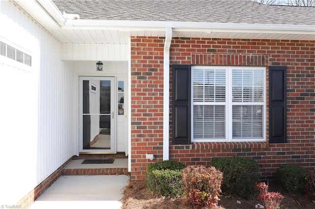 entrance to property featuring visible vents, brick siding, and roof with shingles