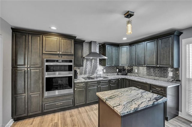 kitchen with double oven, a center island, light stone countertops, black electric cooktop, and wall chimney exhaust hood