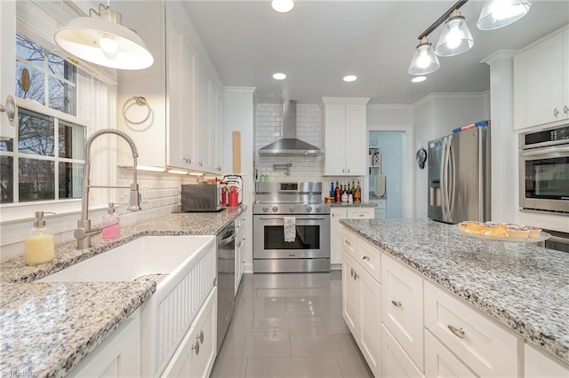 kitchen with wall chimney range hood, appliances with stainless steel finishes, light stone counters, white cabinets, and decorative light fixtures