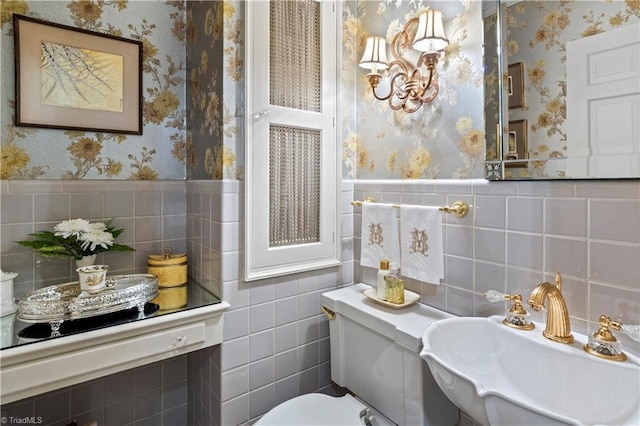bathroom with wallpapered walls, a sink, tile walls, toilet, and a wainscoted wall