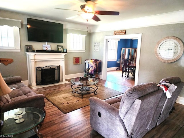 living room featuring ceiling fan, wood finished floors, a fireplace with flush hearth, ornamental molding, and baseboards