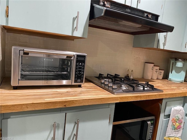 kitchen featuring butcher block countertops, under cabinet range hood, backsplash, stainless steel gas stovetop, and a toaster