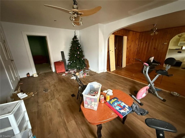 dining room featuring ceiling fan, arched walkways, wood finished floors, baseboards, and wooden walls