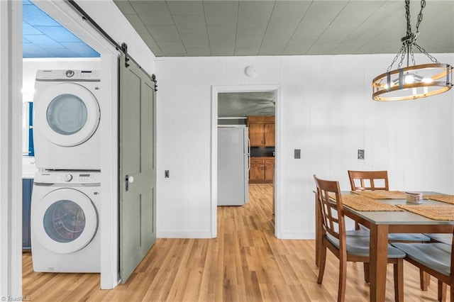 clothes washing area with a notable chandelier, stacked washer / drying machine, a barn door, light wood-style floors, and laundry area